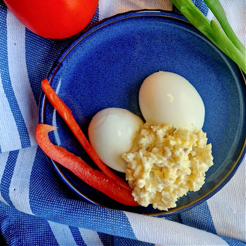 Niebieski talerzyk z dwiema połówkami jajka, na których ułożony jest sos tatarski. Obok ułożone są paski czerwonej papryki, pomidor i zielenina. Całość ustawiona na biało-niebieskiej ściereczce.