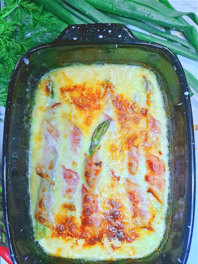 Czarne naczynie żaroodporne wypełnione upieczonymi szparagami owiniętymi w boczek. Danie jest lekko przyrumienione, widać złocistą zapieczoną powierzchnię. Obok naczynia na białym blacie leży cebula dymka i natka pietruszki.