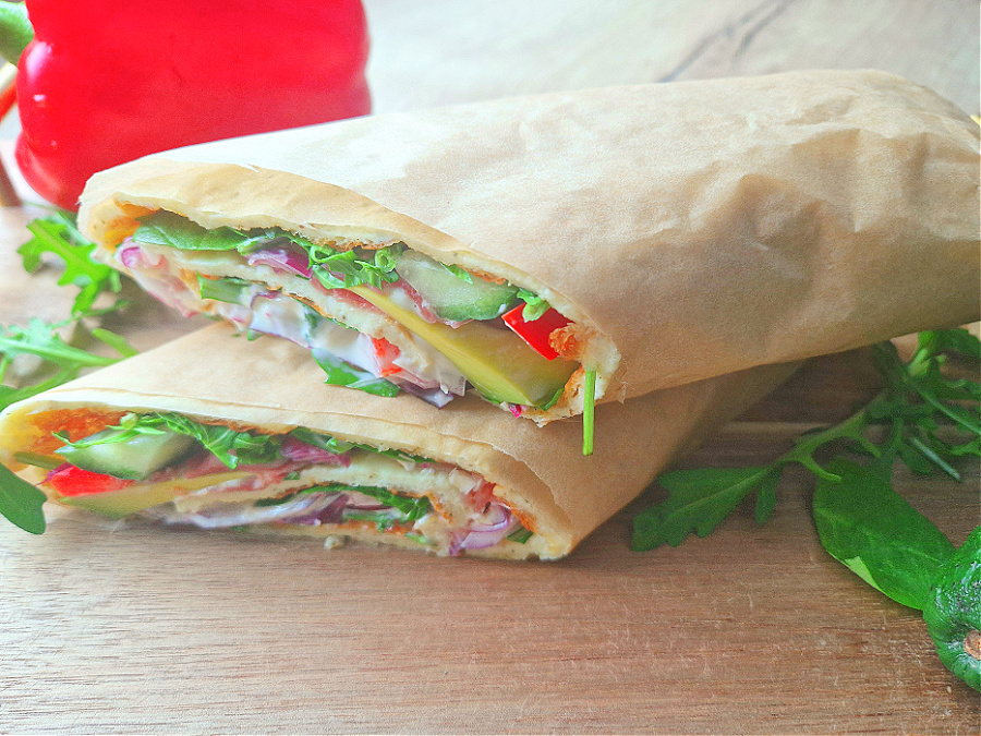 Ujęcie z boku. Wrap z serka wiejskiego w przekroju. Cienkie, elastyczne ciasto widać od środka, a w nadzieniu kolorowe warzywa i szynka. Wrap leży na drewnianym blacie, obok liście rukoli i czerwona papryka.