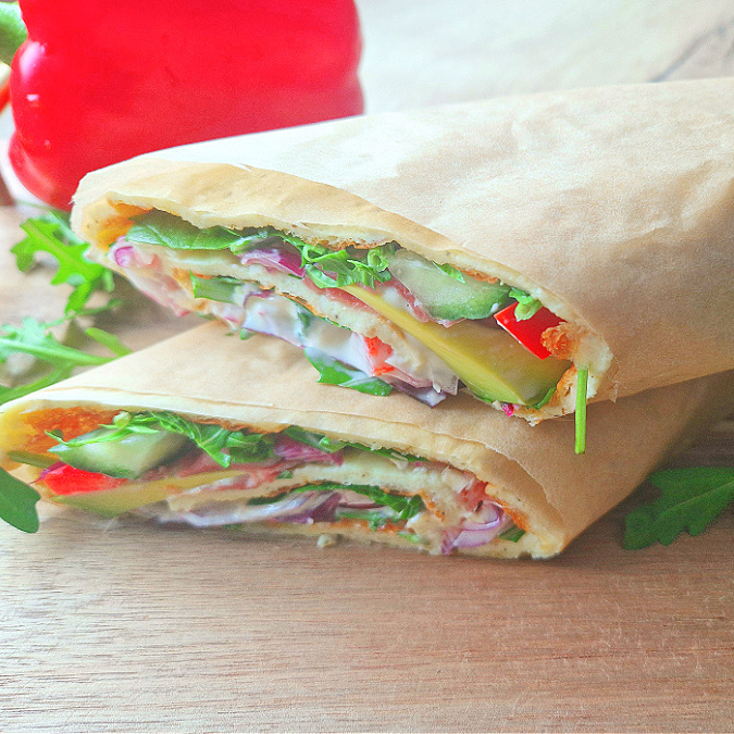 Kwadratowe zdjęcie główne. Wrap z serka wiejskiego, zawinięty w papier do pieczenia. W środku widoczne kolorowe nadzienie: warzywa i szynka. Obok świeża rukola i czerwona papryka.