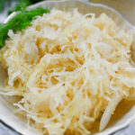 Kwadratowe zdjęcie przedstawiające miseczkę z kapustą kiszoną i gałązką świeżego koperku obok, na jasnym tle.