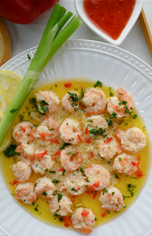 Eleganckie podanie krewetek po hiszpańsku w maśle i czosnku na białym talerzu, z trzema plasterkami cytryny i kawałkiem cebulki dymki. Krewetki posypane zieloną pietruszką i czerwoną papryką, w tle biała miseczka z sosem i pałeczki.