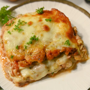 Kwadratowy kawałek keto lasagne z cukinii posypany natką pietruszki, ułożony na biało-złotym talerzyku. Tło jasne, neutralne.