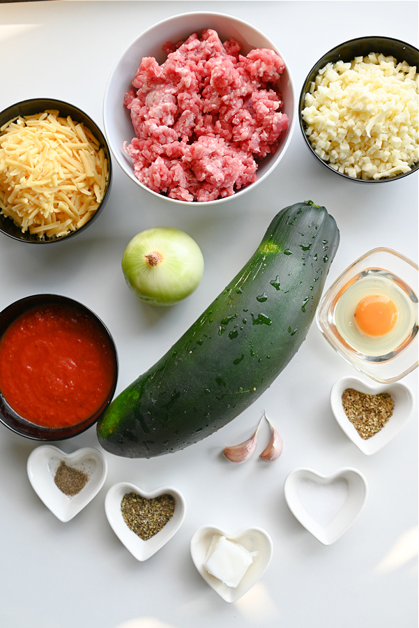 Na białym blacie ułożone składniki do przygotowania keto lasagne z cukinii: cukinia, mięso mielone w miseczce, ser mozzarella, ser cheddar, śmietanka 30%, passata pomidorowa bez cukru, czosnek, cebula, jajko, oliwa, sól, pieprz, oregano i bazylia.