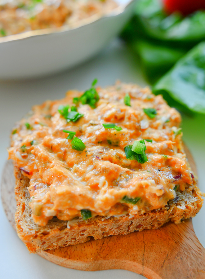 Zbliżenie na kromkę chleba posmarowaną pastą ze śledzia w puszce i posypaną szczypiorkiem, ułożoną na małej drewnianej deseczce na białym blacie. W tle widać miskę z pastą i zielone liście sałaty.