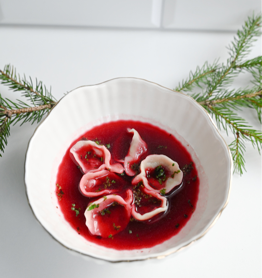 Biała miseczka wypełniona klarownym czerwonym barszczem, w którym pływają jasne uszka z grzybami, ustawiona na jasnym tle z delikatnymi gałązkami choinki obok.