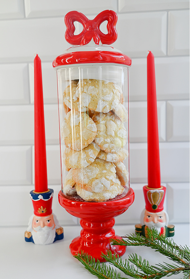 Ciasteczka cytrynowe popękane w świątecznym pojemniku i z świątecznymi świecznikami na białym blacie na tle białych kafelek.