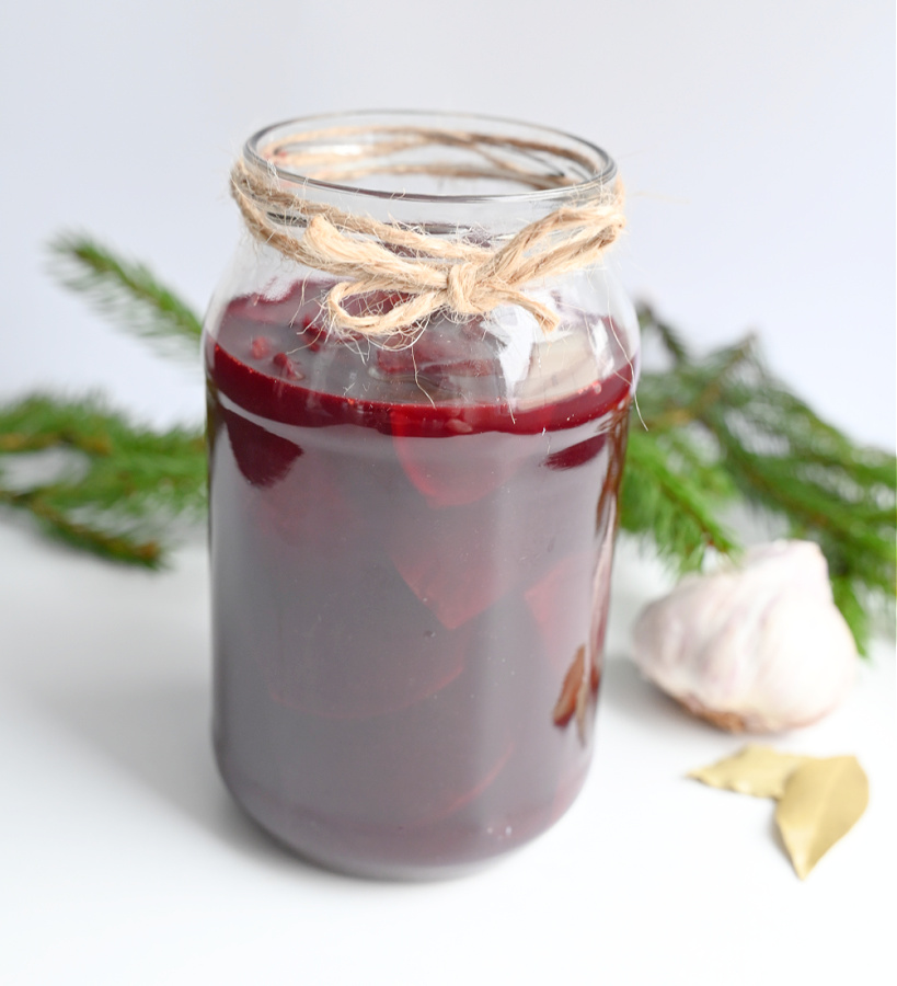 Słoik ze sfermentowanym zakwasem z buraków owinięty sznurkiem tuż pod zakrętką. W środku intensywnie czerwony płyn i plasterki buraka. Obok leży liść laurowy, główka czosnku i zielone gałązki choinki na białym tle