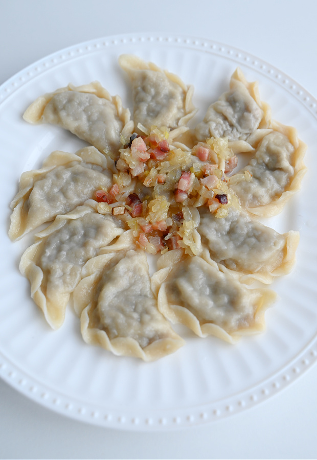 Ugotowane pierogi z białą kaszanką ułożone na białym talerzu, na środku porcja okrasy z boczku i cebuli, całość na jasnym blacie