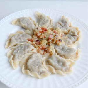 Pierogi z białą kaszanką podane na białym talerzu, na środku okrasa z cebuli i boczku, jasne tło