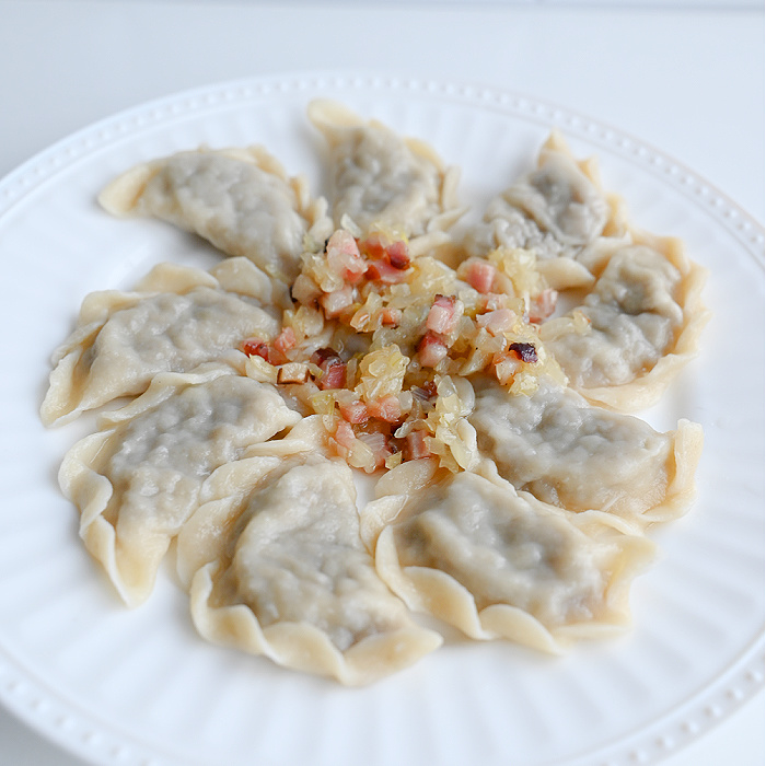 Pierogi z białą kaszanką podane na białym talerzu, na środku okrasa z cebuli i boczku, jasne tło