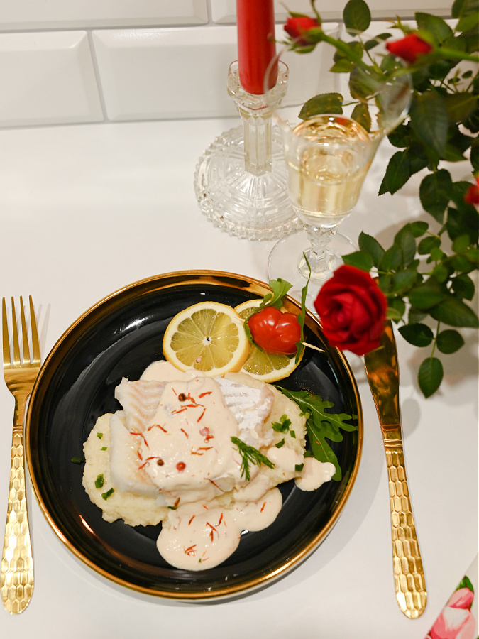 Romantyczna kolacja z dorszem w sosie z różowego pieprzu, czerwona świeca, kieliszek białego wina, róża i czarny talerz ze złotą obwódką