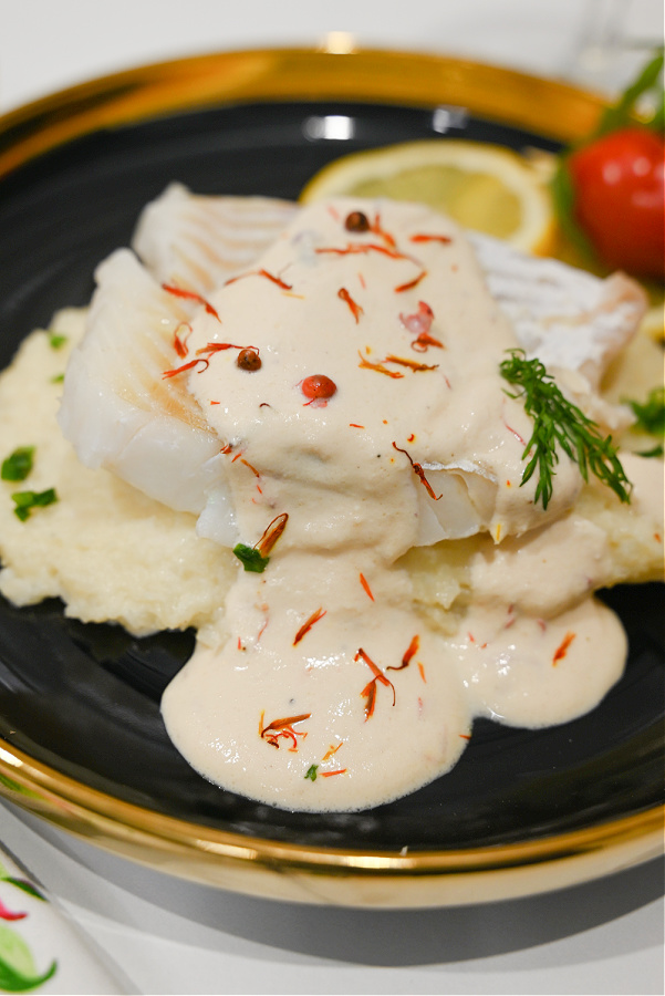 Dorsz na puree z kalafiora z sosem z różowego pieprzu i szafranu, czarny talerz ze złotą obwódką, ujęcie z boku na białym blacie