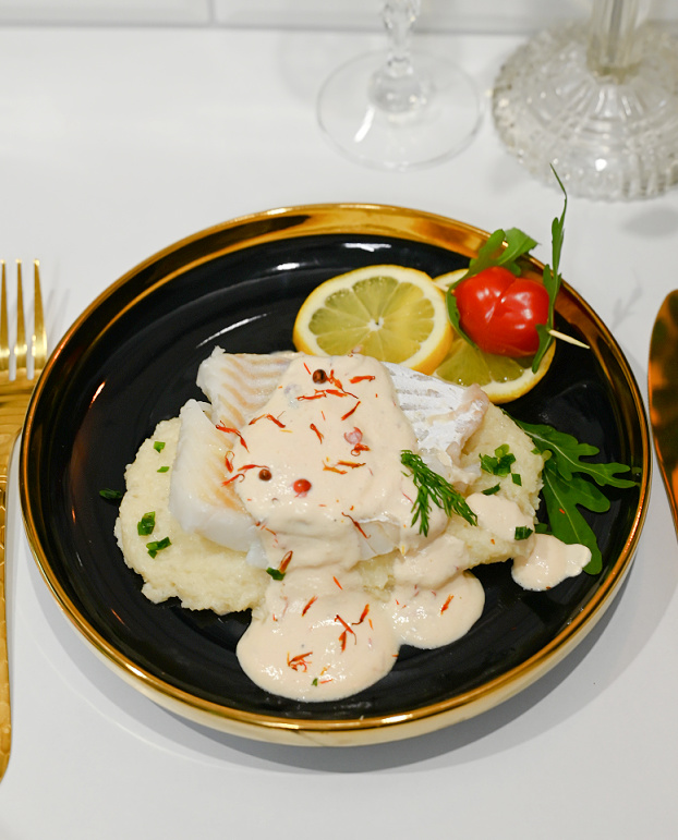 Widok z góry na dorsza w sosie z różowego pieprzu na puree z kalafiora, czarny talerz ze złotą obwódką, złote sztućce