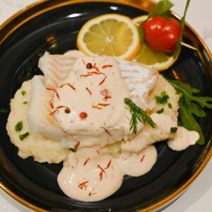 Dorsz w sosie z różowego pieprzu podany na puree z kalafiora z plasterkiem cytryny i sercem z pomidorka, czarny talerz ze złotą obwódką na białym blacie