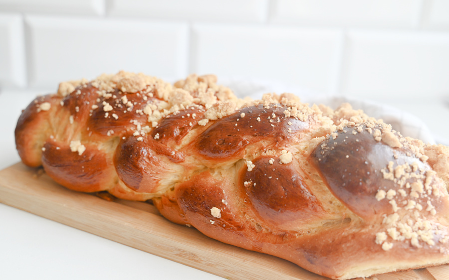 Cała podłużna chałka z kruszonką na drewnianej desce ustawiona na tle białych kuchennych kafelków.