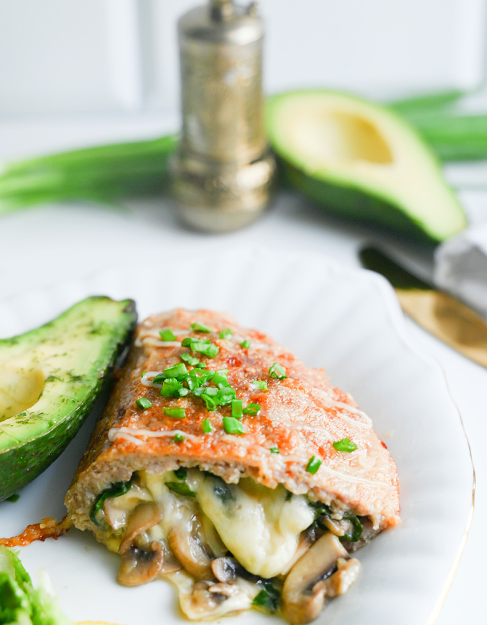Pojedyncza, upieczona kieszonka o mocno zrumienionej, grudkowatej powierzchni, która leży na trzech soczyście zielonych liściach szpinaku na białym talerzu. W rozmytym tle widać połówkę zielonego awokado i srebrny młynek do pieprzu.