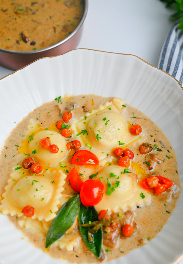 Ravioli podane na białym talerzu w kremowym sosie grzybowym, udekorowane pomidorkami koktajlowymi, zielonymi liśćmi szałwii i posiekaną natką pietruszki