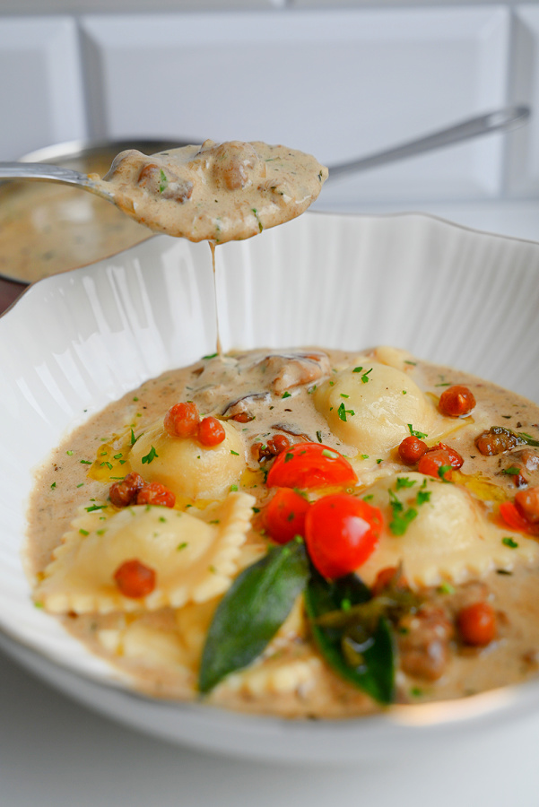 Zbliżenie na gorącą, białą miskę pełną ravioli z gęstym, kremowym sosem grzybowym. Nad miską srebrna łyżka unosi solidną porcję sosu ze śmietanki i podgrzybków. Danie udekorowane jest połówkami pomidorków koktajlowych, chrupiącymi kuleczkami, świeżą pietruszką i smażoną szałwią. W tle widać patelnię.
