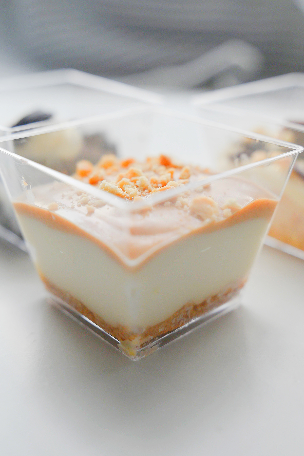 Mini deserek serniczek Lotus w przezroczystym, kwadratowym pucharku. Widoczna jasna masa serowa, gładka, jasnobrązowa polewa karmelowa oraz posypka z pokruszonych ciasteczek na wierzchu. W rozmazanym tle widać dwa inne deserki.
