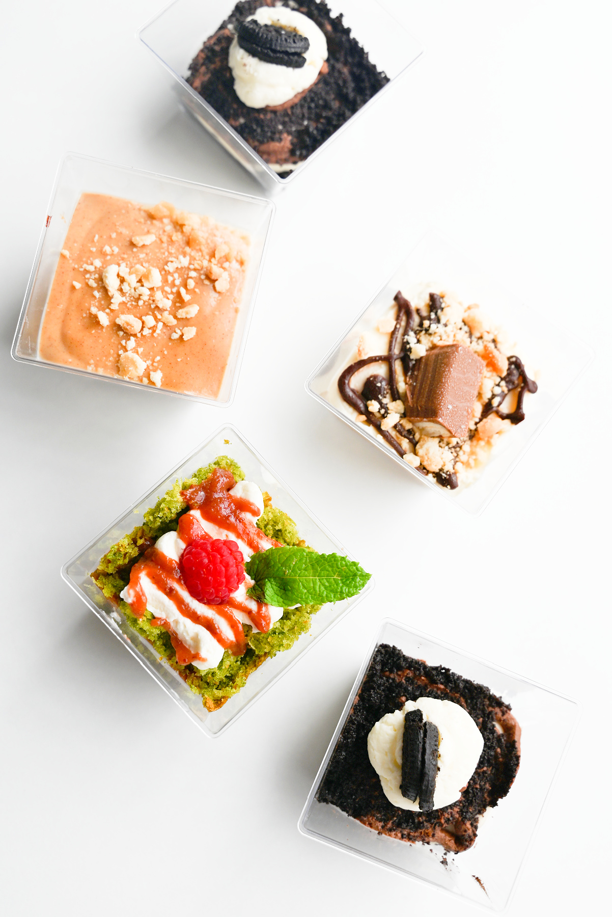 Pięć różnych mini deserków w kwadratowych pucharkach, sfotografowanych z góry na czystym, białym tle. Wśród nich serniczek Lotus z karmelową polewą, deserek z zielonym ciastem i maliną oraz deserki z ciasteczkami Oreo i czekoladą.