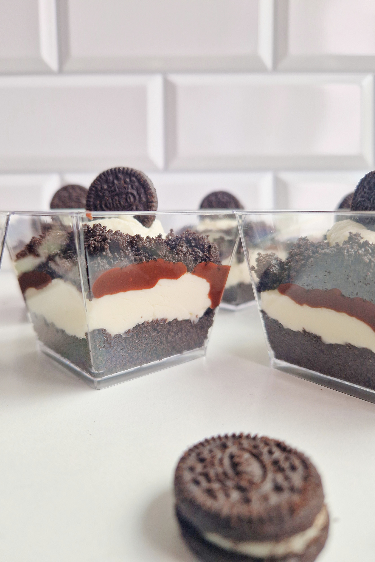 Gotowe deserki Oreo w kwadratowych pucharkach na tle jasnej ściany z kafelków. Widoczne idealne warstwy i dekoracja z ciastka.