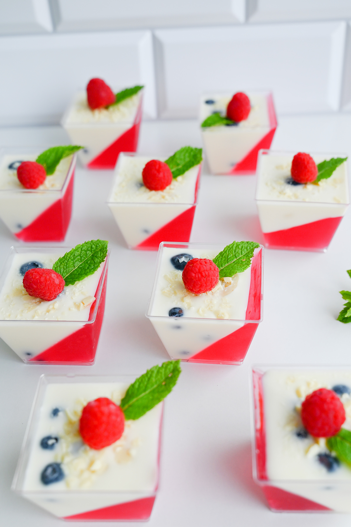 Rzędy kwadratowych słoiczków z panna cottą i galaretką pod skosem, widziane z góry na białym tle.