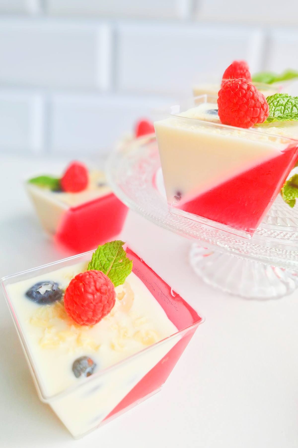 Mini deserki panna cotta z czerwoną galaretką pod skosem, udekorowane świeżą maliną, borówkami i miętą na jasnym tle.