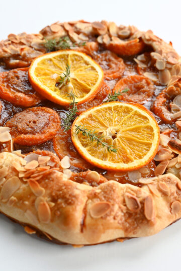 Pieczona rustykalna galette z morelami, plastrami pomarańczy i płatkami migdałów na białym tle.