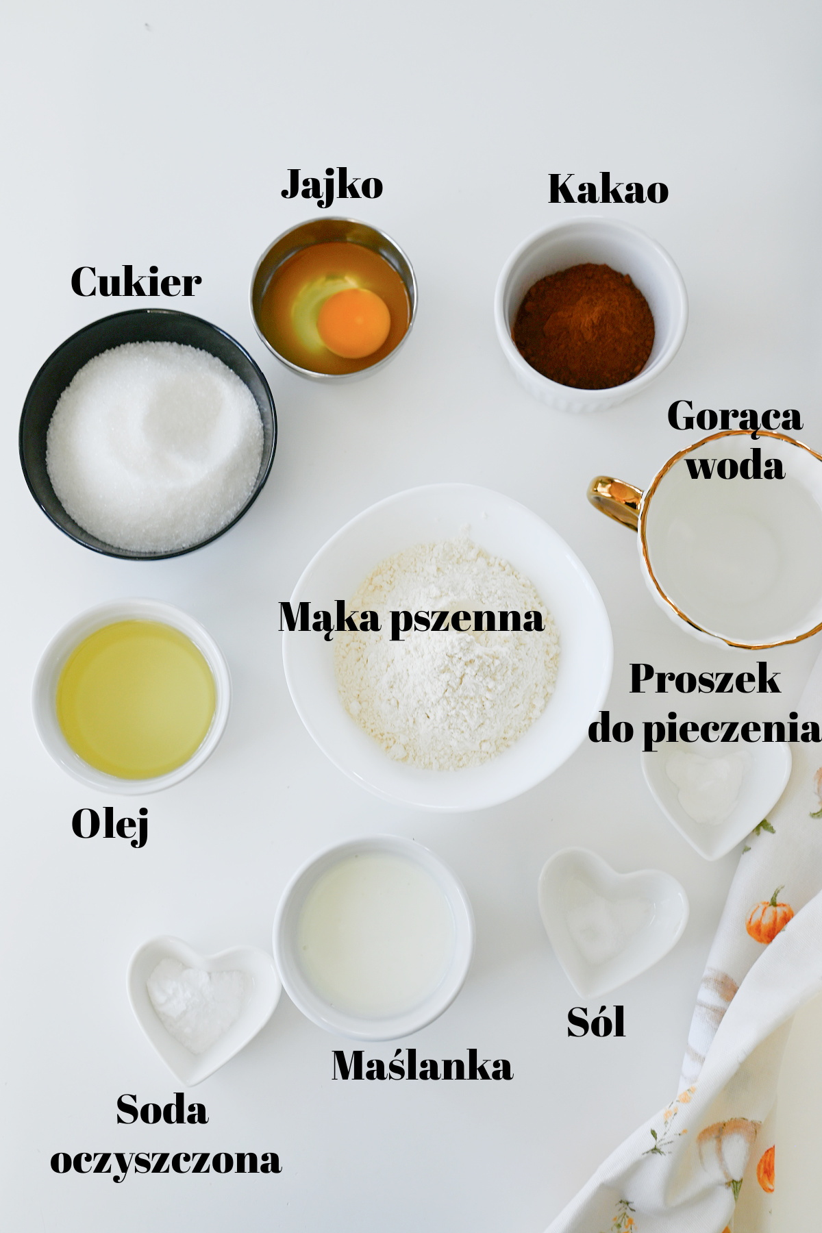 Składniki do ciasta czekoladowego w białych i czarnych miseczkach na jasnym blacie z czarnymi podpisami.